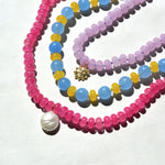 Pink agate and wonky pearl necklace in sunlight beside blue, yellow and lilac granny bead necklaces