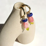 Pair of lapis lazuli, apricot thulite and Korean peridot charms on gold hoops, hung on a ceramic vase in sunlight