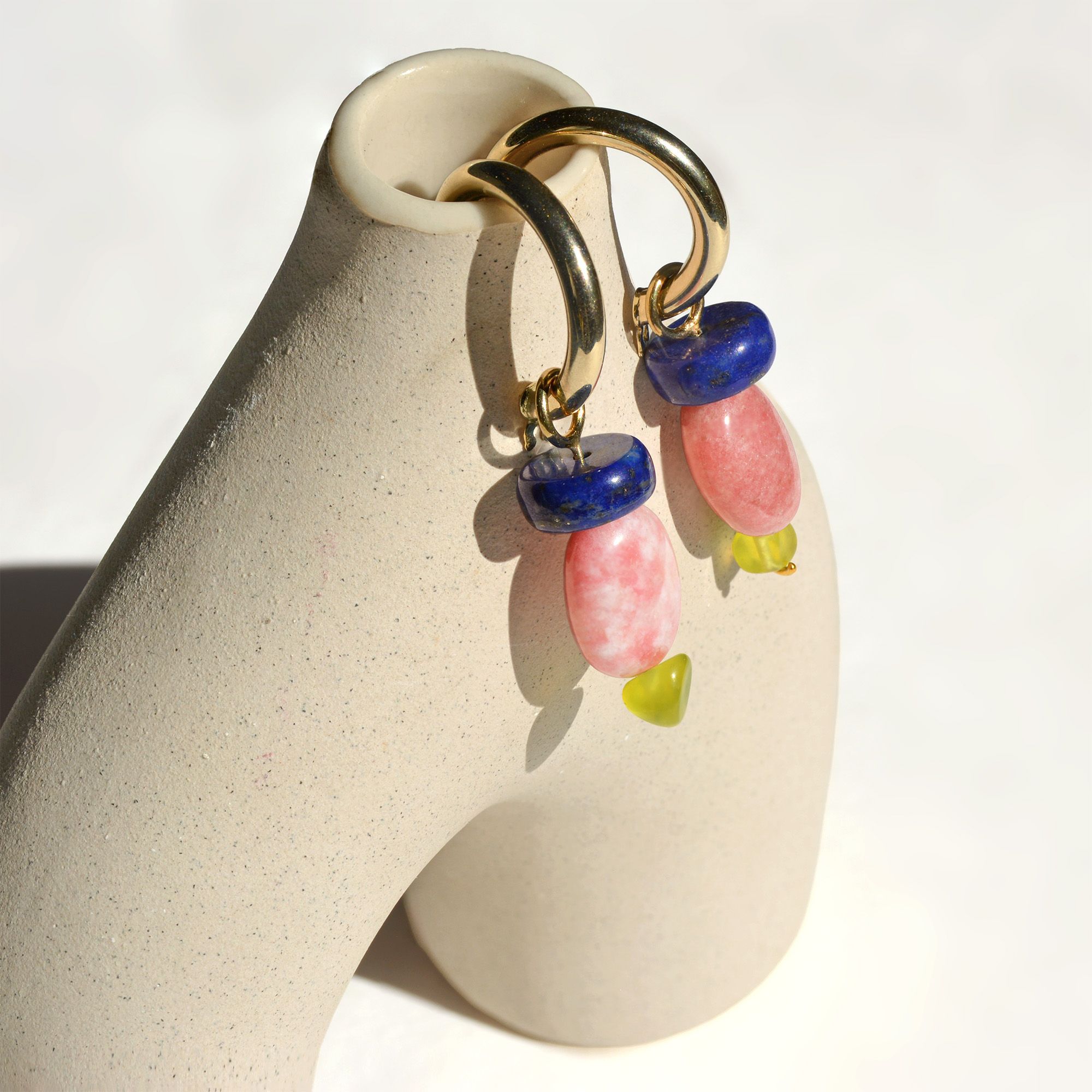 Pair of lapis lazuli, apricot thulite and Korean peridot charms on gold hoops, hung on a ceramic vase in sunlight