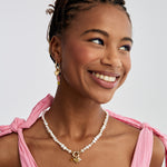 A smiling woman wearing a white pearl necklace with gold pendants and a charms on a hoop earring, styled with a pink tie-neck top against a light gray background.