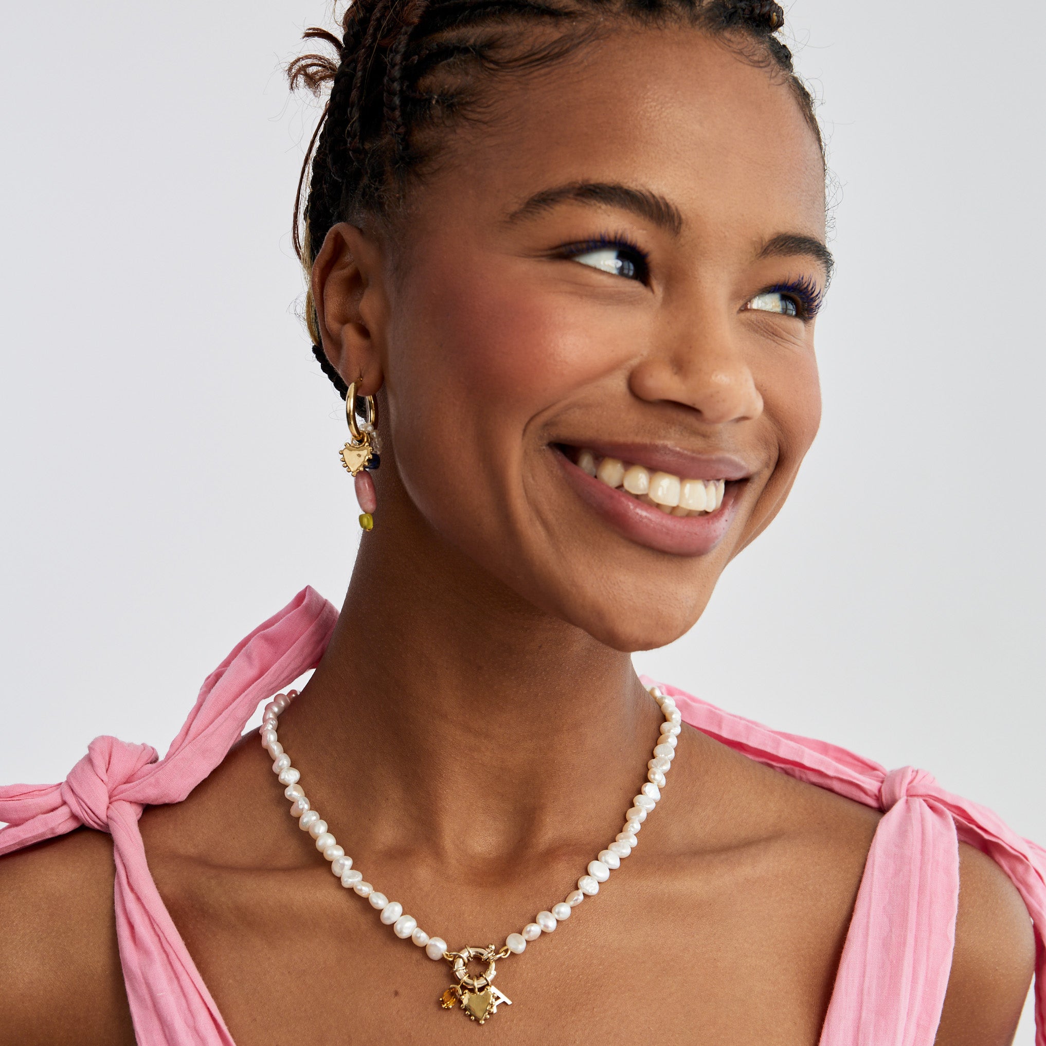 A smiling woman wearing a white pearl necklace with gold pendants and a charms on a hoop earring, styled with a pink tie-neck top against a light gray background.