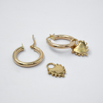 Two gold earrings arranged on a plain light background: one hoop with a dangling heart charm, and another hoop earring with a separate heart charm piece.