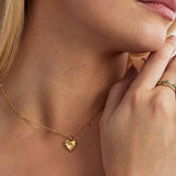 A close-up lifestyle shot of a model wearing a delicate gold chain necklace with a small, textured gold heart pendant. The model's neck and hand are visible, with a gold ring also shown on her finger.
