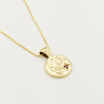 Gold necklace with a round pendant featuring a face design and a small red gemstone on a light gray background.