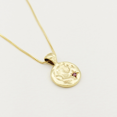 Gold necklace with a round pendant featuring a face design and a small red gemstone on a light gray background.