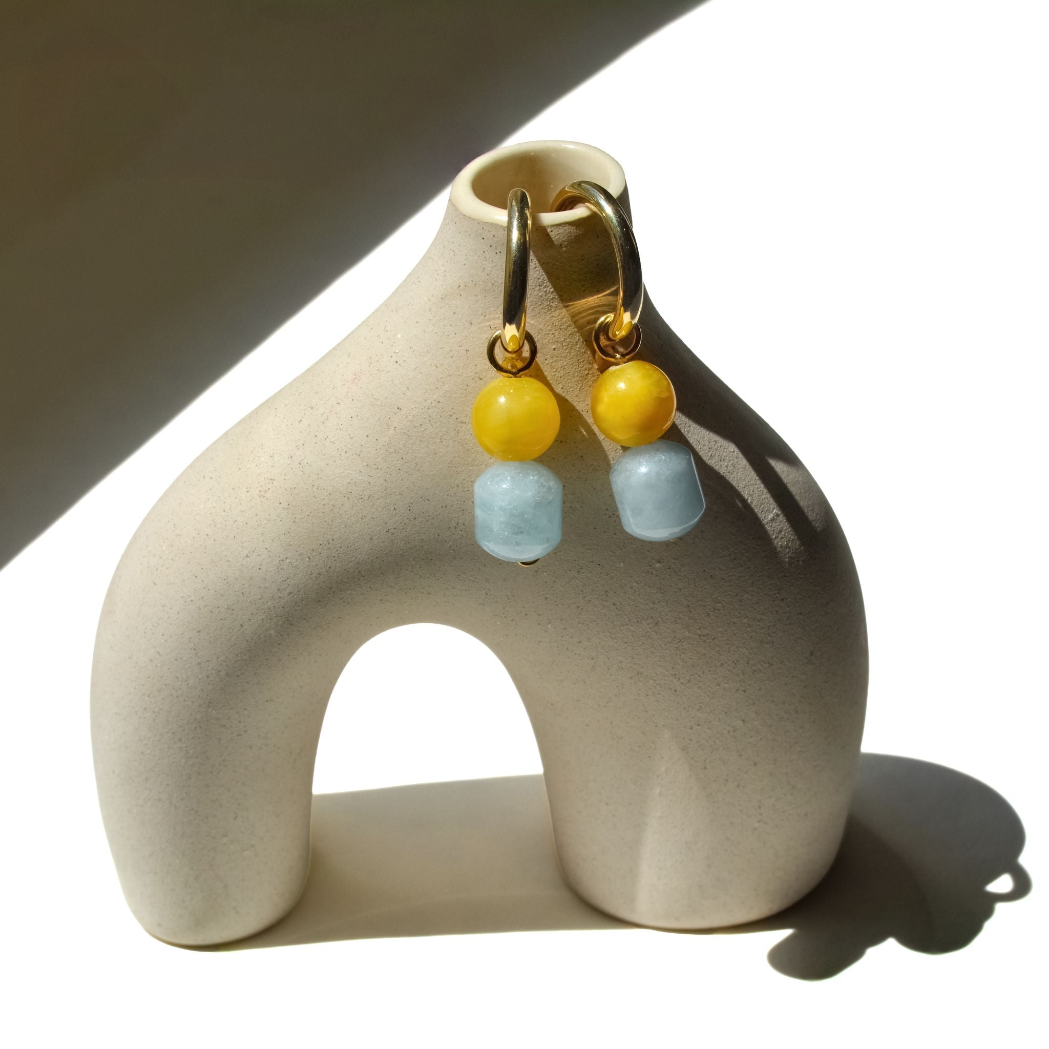 Pair of mango jade and aquamarine charms on gold hoops, hung on a ceramic arch vase in sunlight