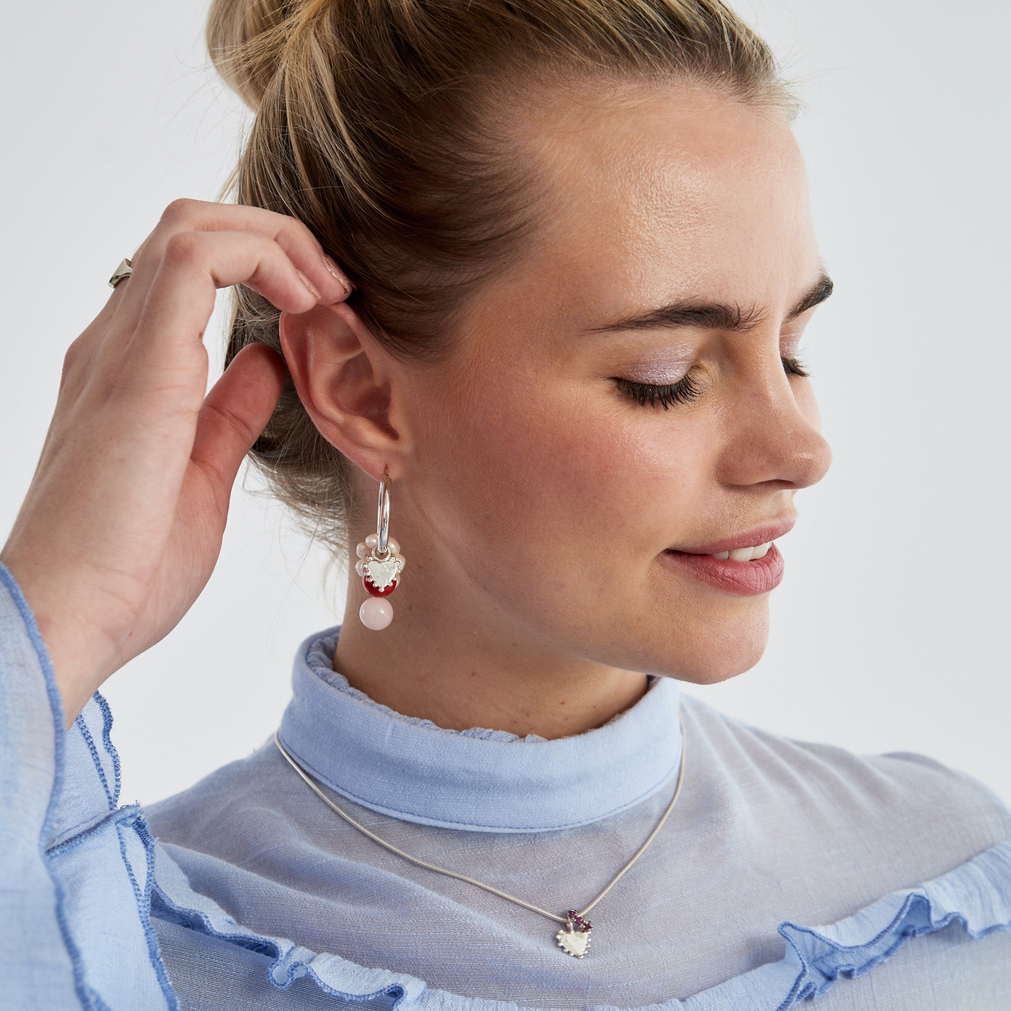A woman with blonde hair in a bun models a silver dangle earring and matching necklace. The jewelry features a white heart-shaped charm with red accents and a light pink bead.