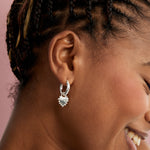 A close-up side profile of a woman wearing a silver hoop earring with a dangling heart-shaped silver charm, set against a solid pink background.
