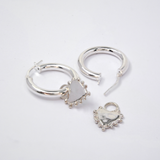 Two silver earrings arranged on a plain light background: one hoop with a dangling heart charm, and another hoop earring with a separate heart charm piece.