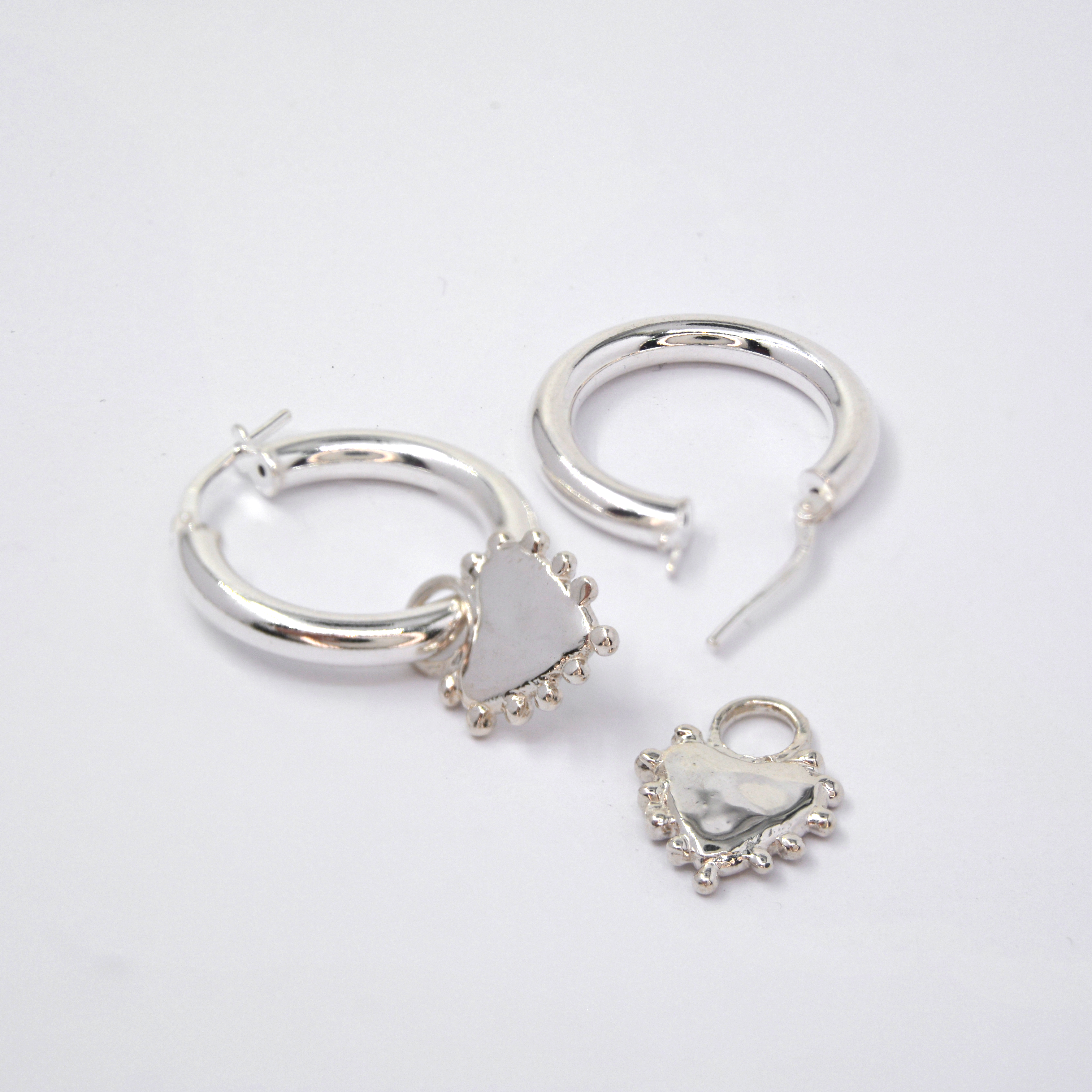 Two silver earrings arranged on a plain light background: one hoop with a dangling heart charm, and another hoop earring with a separate heart charm piece.