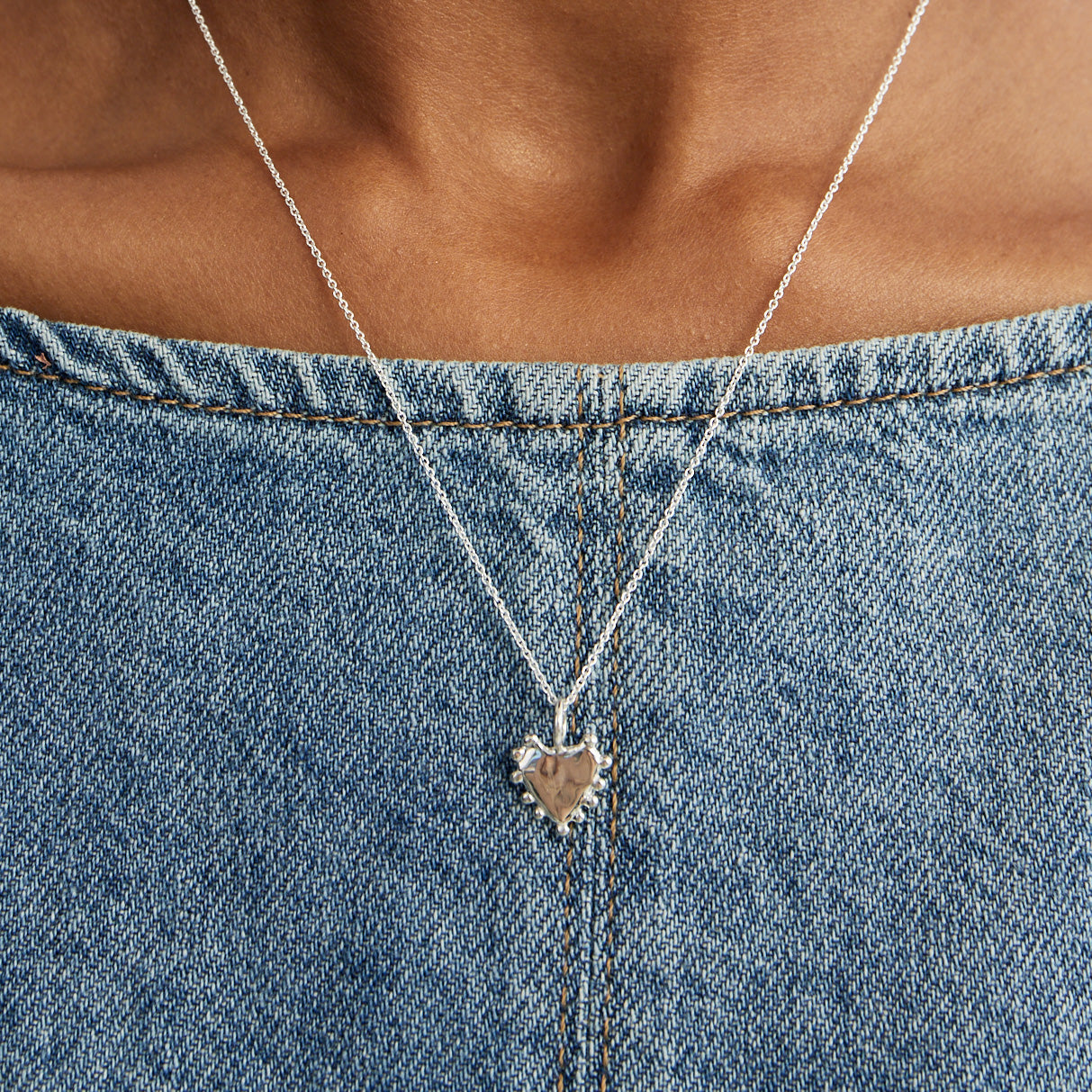 A small silver heart-shaped pendant charm with a beaded border around it's edges on a chain on a plain light gray background. styled over a blue denim top.
