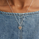 A small silver heart-shaped pendant charm with a beaded border around it's edges on a chain on a plain light gray background. styled over a blue denim top.
