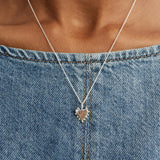 A small silver heart-shaped pendant charm with a beaded border around it's edges on a chain on a plain light gray background. styled over a blue denim top.