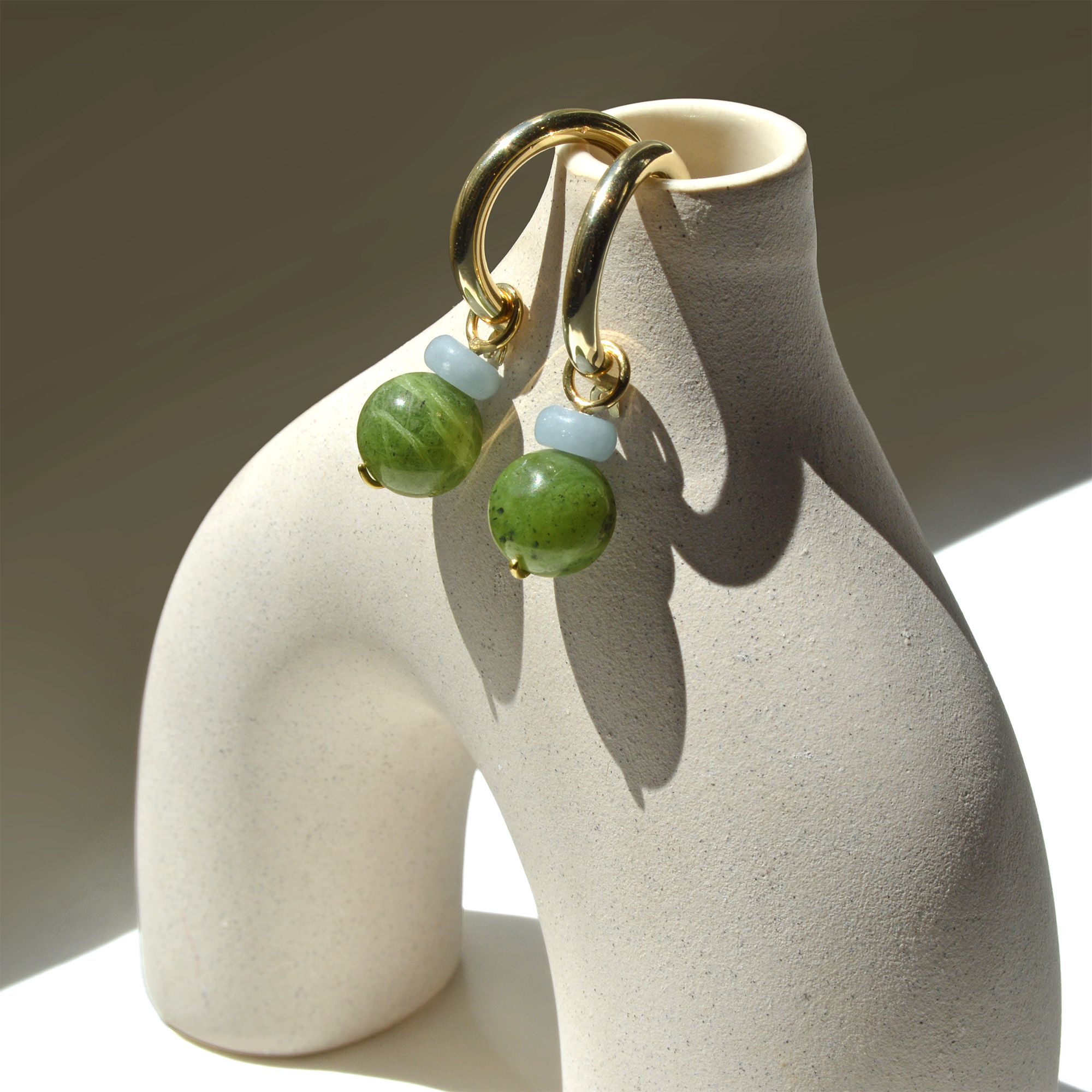 Pair of angelite and Canada jade charms on gold hoops, hung on a ceramic vase in sunlight
